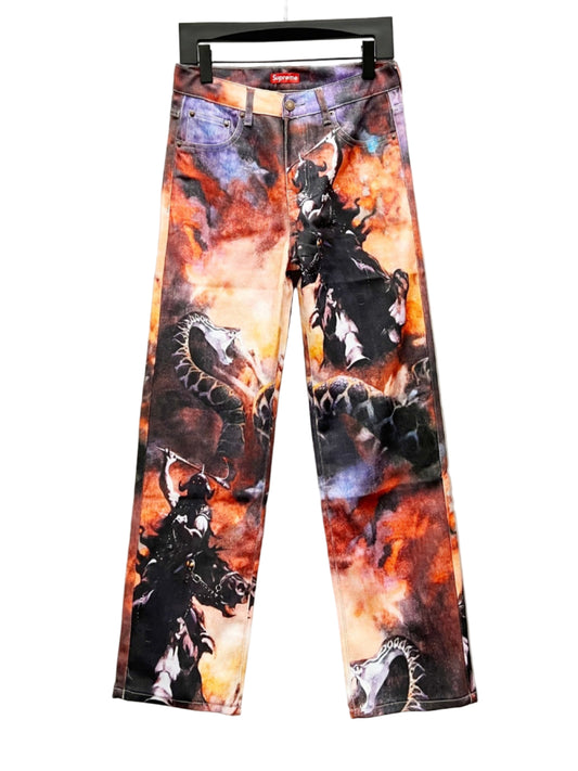 Supreme Firestorm graphic denim pants with bold orange, black, and grey abstract print, displayed on a hanger against a light grey wall.